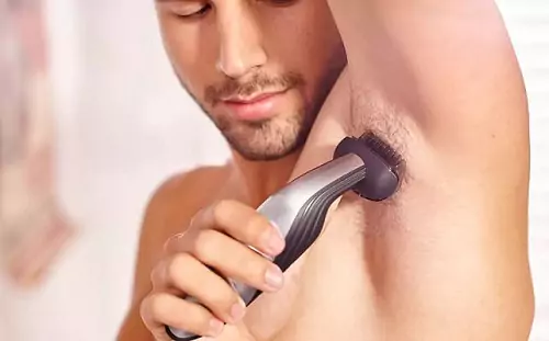Homem branco sem camisa utilizando aparador de pelos para depilar as axilas.
