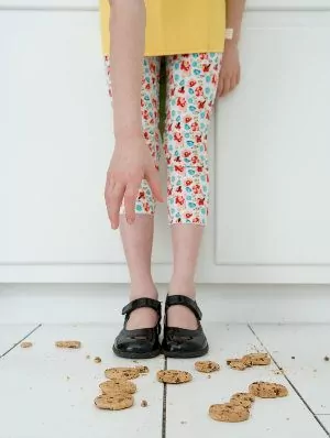 Menina se abaixando para limpar biscoitos espalhados pelo chão.