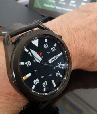 Foco no Galaxy Watch 3 preso no pulso de homem, com um mostrador de relógio analógico como fundo de tela.