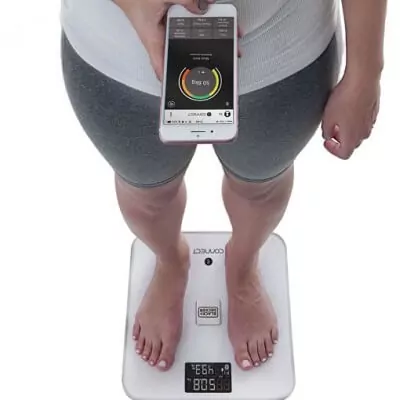 Mulher em cima de balança inteligente branca conferindo sua composição corporal no smartphone.