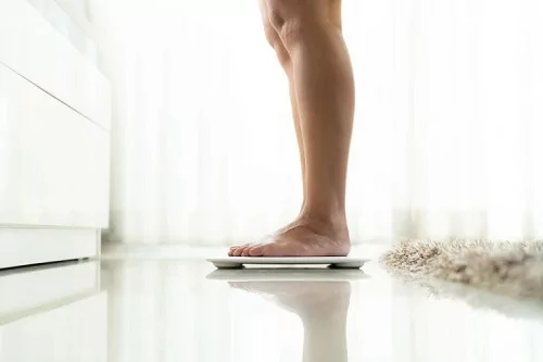 Vista lateral das pernas de uma pessoa em cima de uma balança, em um piso nivelado.
