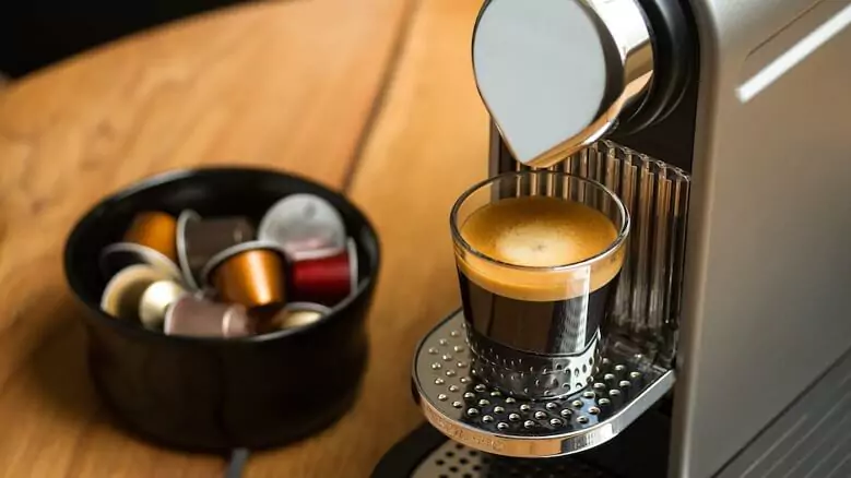 Abertura do post sobre a melhor cafeteira expresso: Cafeteira cinza com um copo de café na bandeja, ao lado de cápsulas.