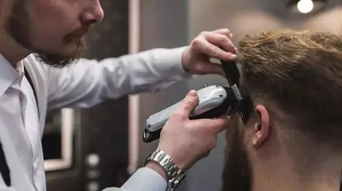 Vista lateral de barbeiro usando cortador de cabelo sem fio para cortar a lateral do cabelo de cliente.
