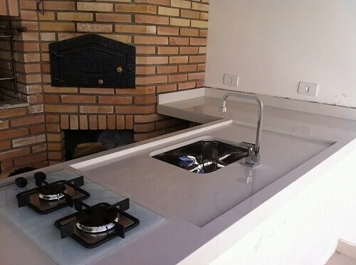 Cooktop de duas bocas de mesa de vidro cor gelo instalado em balcão branco com churrasqueira de tijolos ao fundo.