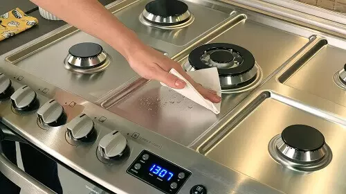 Mão feminina limpando fogão 5 bocas mesa de inox com um pano húmido.